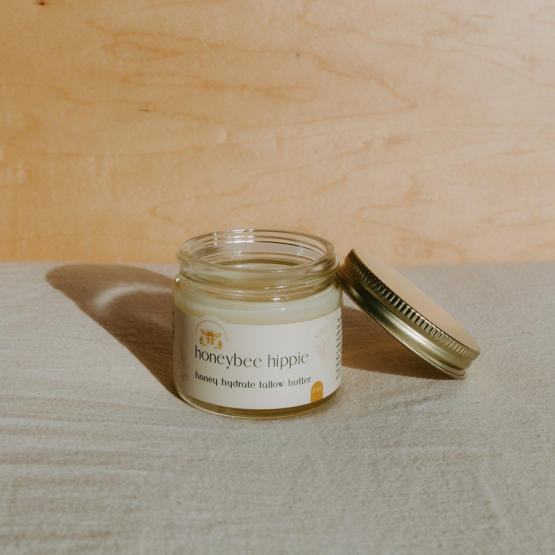 An open jar of Honeybee Hippie's honey hydrate tallow butter, infused with organic herbs, sits on a soft surface. Sunlight and a warm wooden backdrop cast gentle shadows.