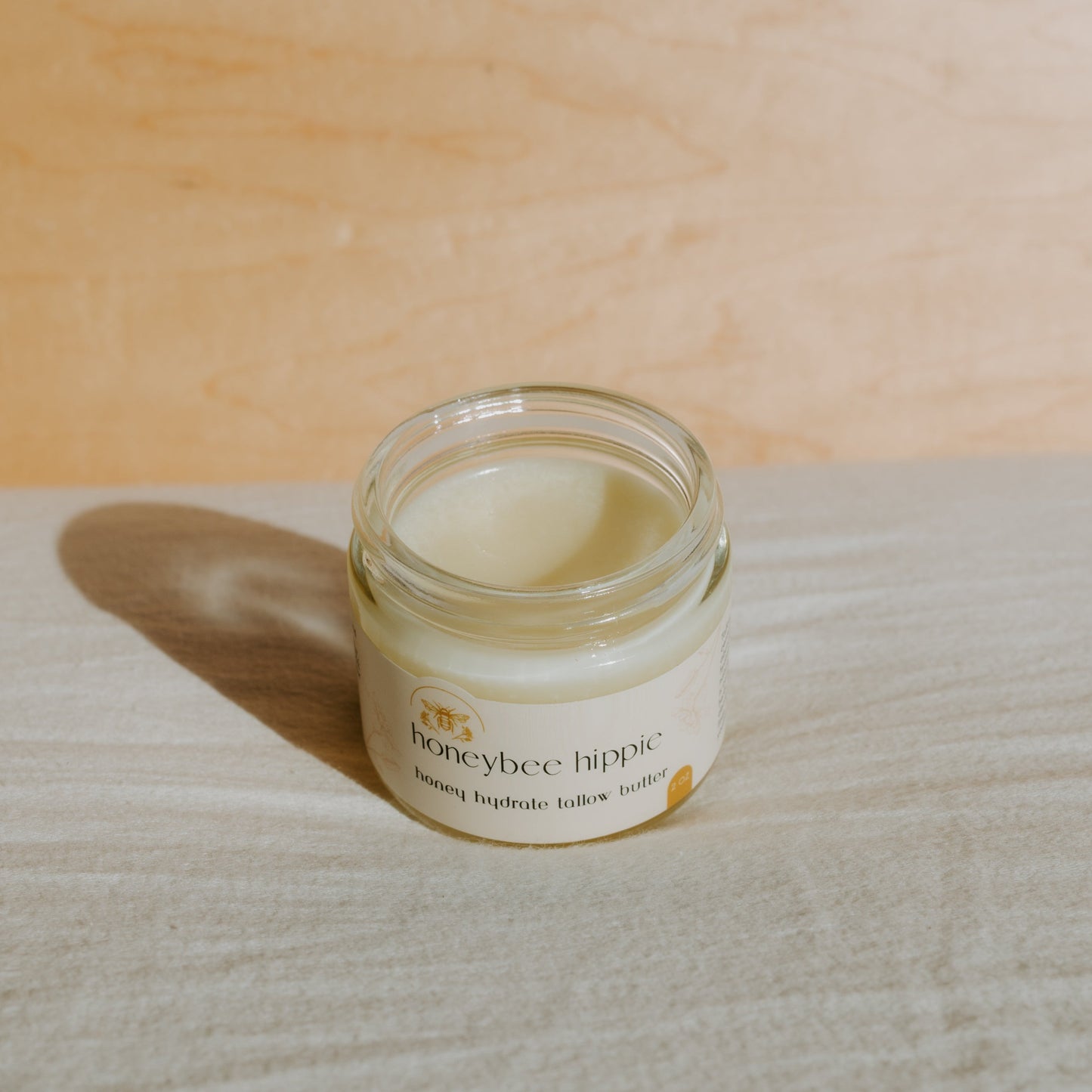 A Honeybee Hippie glass jar of "honey hydrate tallow butter" with Manuka honey hints rests on a textured surface. The softly lit background adds a warm, natural ambiance.