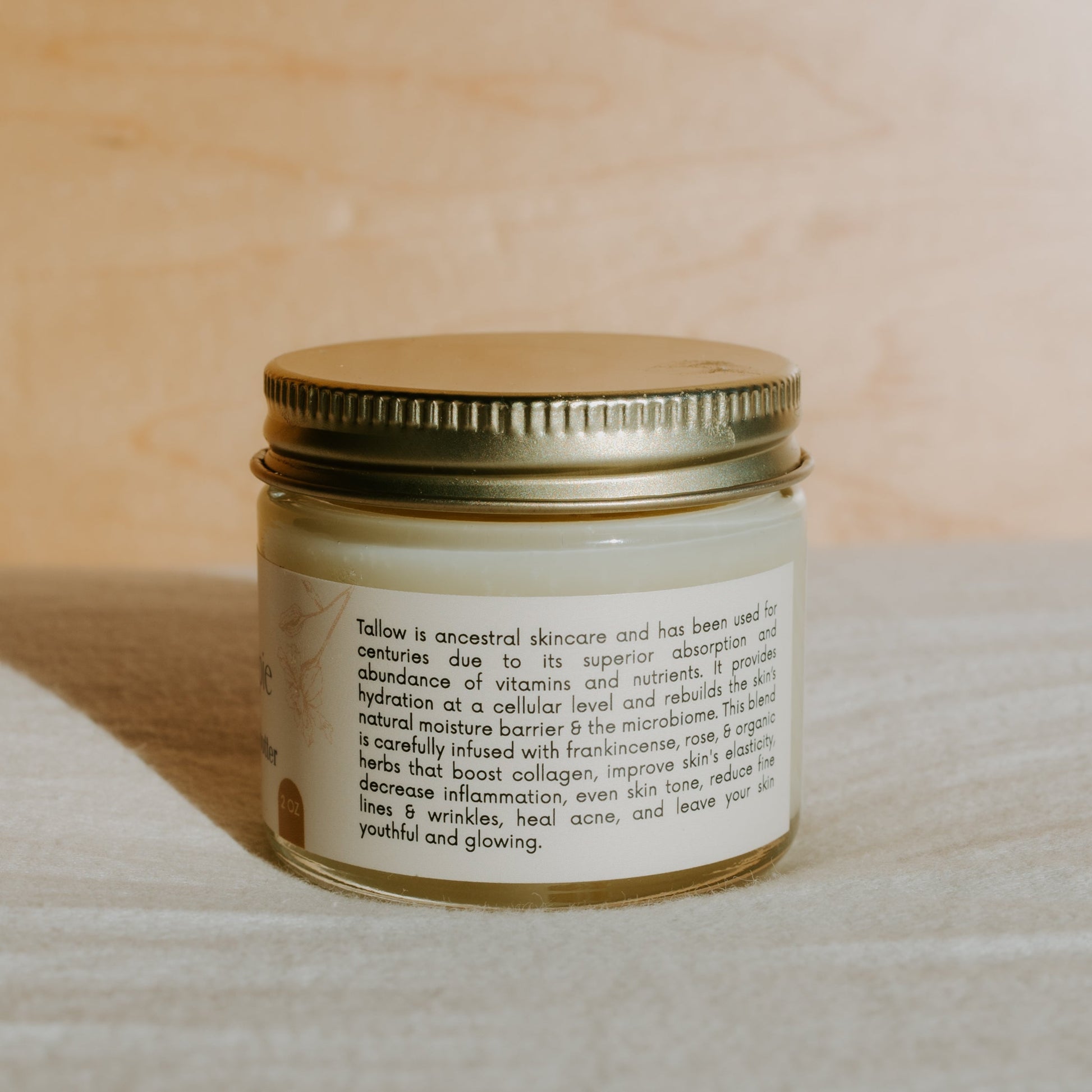 A small jar of Honeybee Hippie's honey hydrate tallow butter with a gold lid sits on a light surface. The label highlights benefits like skin hydration, anti-aging, and acne healing. Organic herbs enhance the formula, while a softly blurred background creates a warm and natural aesthetic.