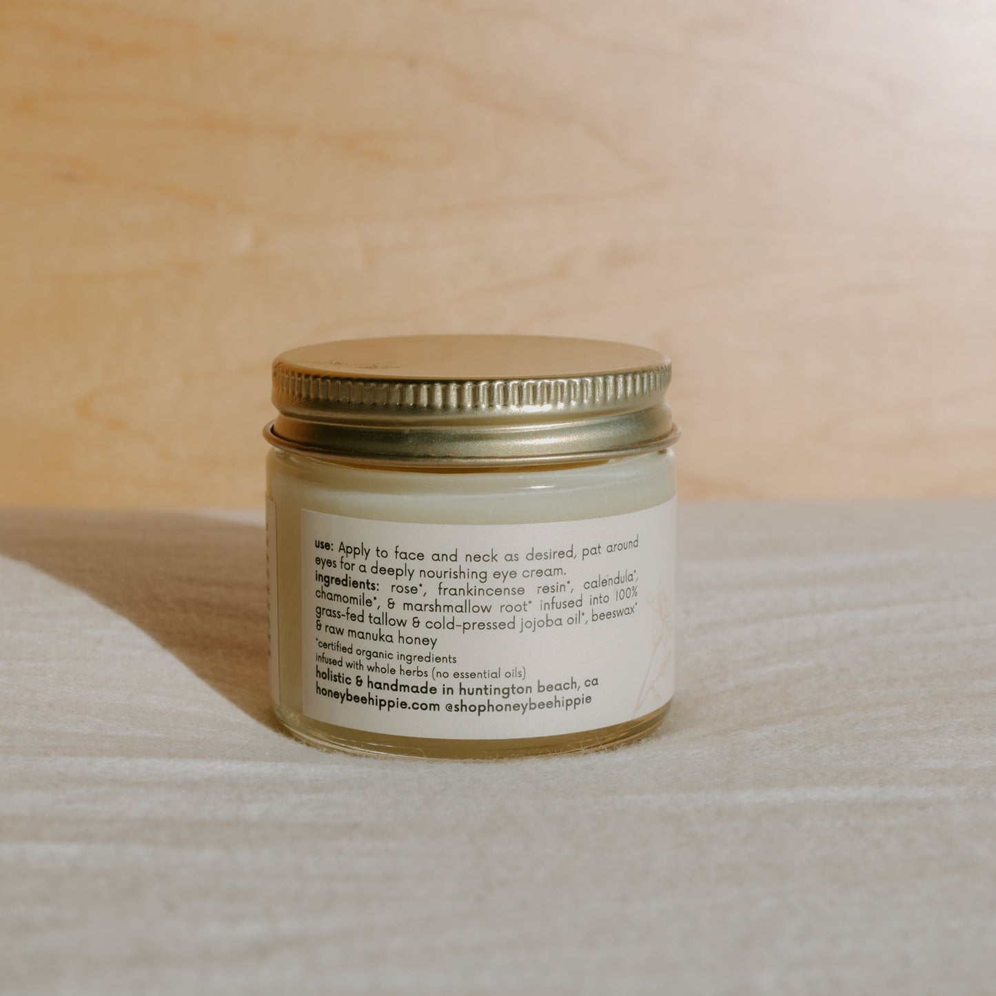 A glass jar with a gold lid beautifully stands against a wooden background. The label of Honeybee Hippie's Honey Hydrate Tallow Butter showcases its rich blend of organic herbs and Manuka honey, providing natural nourishment. Sunlight casts soft shadows, enhancing its allure.