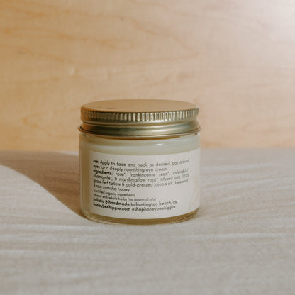 A glass jar with a gold lid beautifully stands against a wooden background. The label of Honeybee Hippie's Honey Hydrate Tallow Butter showcases its rich blend of organic herbs and Manuka honey, providing natural nourishment. Sunlight casts soft shadows, enhancing its allure.