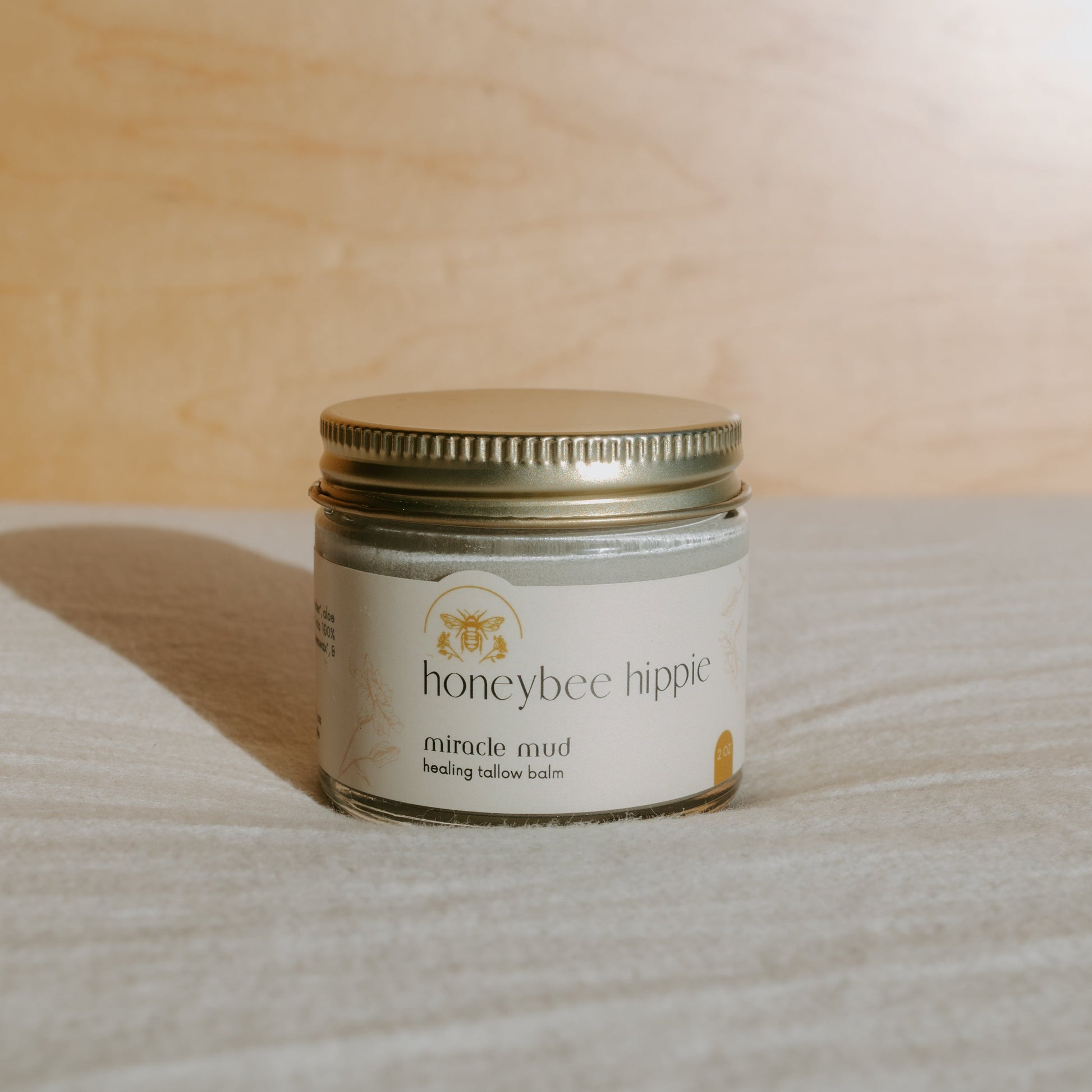 The "miracle mud" by Honeybee Hippie, a tallow-based balm for natural skincare, rests elegantly on light fabric with a wooden background. The small jar features a simple design and metal lid, ideal for soothing dry skin and diaper rash.