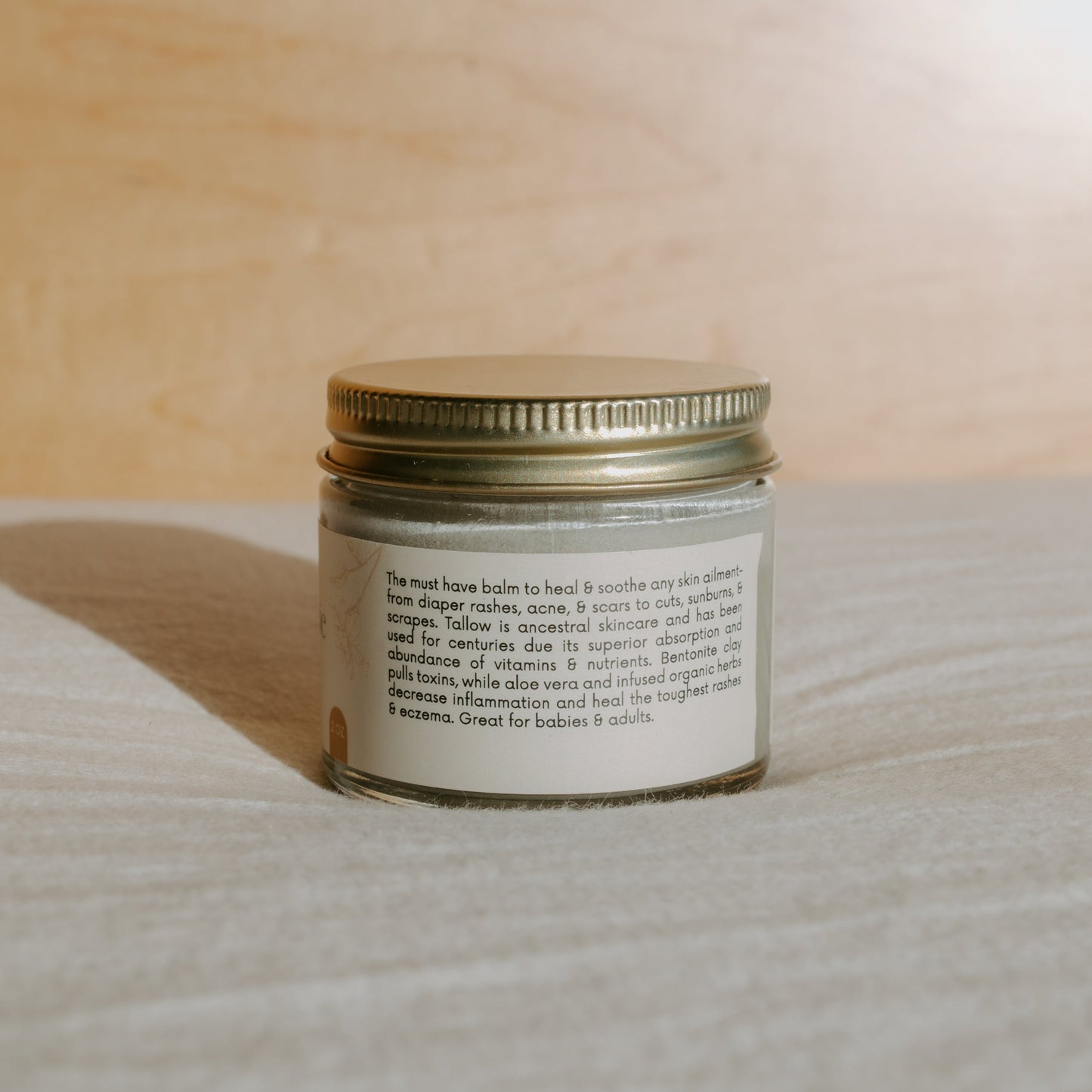 A small jar labeled "Miracle Mud" by Honeybee Hippie, with a metallic lid, sits on a wooden surface in warm sunlight. This tallow-based balm offers soothing relief for skin ailments and natural skincare needs for both babies and adults, including diaper rash relief.