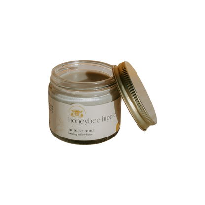 A jar of Honeybee Hippie's "miracle mud" tallow balm, ideal for skincare and diaper rash relief, has a slightly open gold lid. Its label showcases a bee illustration and text, set against a black background.