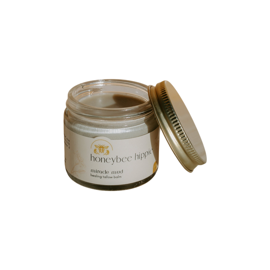 A jar of Honeybee Hippie's "miracle mud" tallow balm, ideal for skincare and diaper rash relief, has a slightly open gold lid. Its label showcases a bee illustration and text, set against a black background.