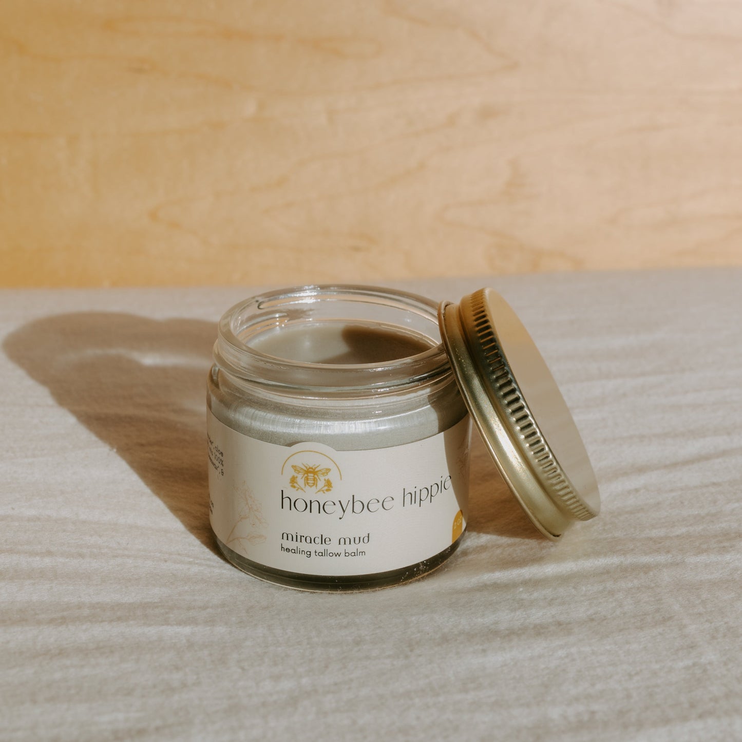 An open glass jar of "Honeybee Hippie Miracle Mud" sits on a textured surface, highlighting its natural skincare benefits. The light label with floral graphics and a bee logo complements the golden lid beside it, casting a shadow—ideal for soothing diaper rash and more.