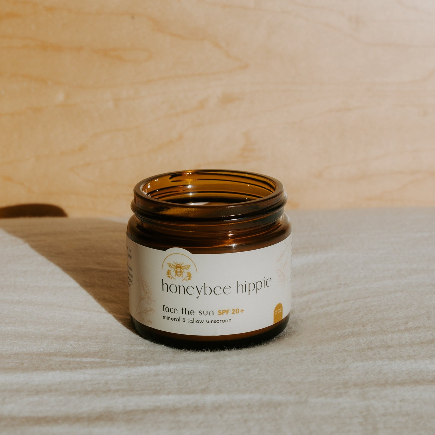 An open brown glass jar labeled “Honeybee Hippie face the sun - herbal tinted mineral & tallow skin protection” sits on light fabric with a wooden background, naturally lit to highlight its UV-blocking, mineral-rich formula.