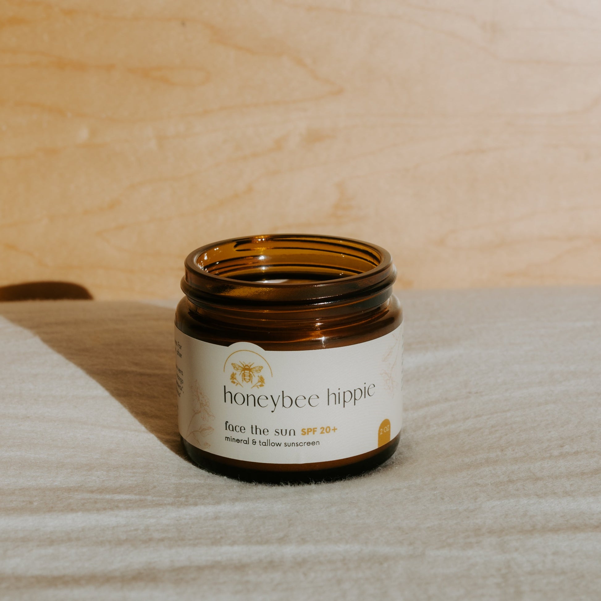An open brown glass jar labeled “Honeybee Hippie face the sun - herbal tinted mineral & tallow skin protection” sits on light fabric with a wooden background, naturally lit to highlight its UV-blocking, mineral-rich formula.