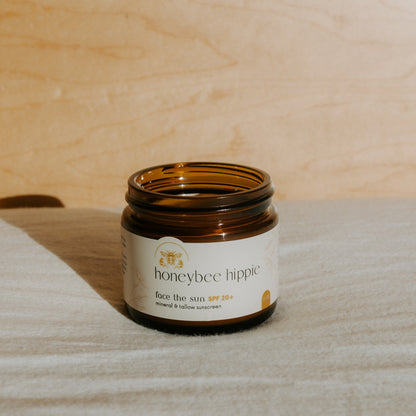 An open brown glass jar labeled “Honeybee Hippie face the sun - herbal tinted mineral & tallow skin protection” sits on light fabric with a wooden background, naturally lit to highlight its UV-blocking, mineral-rich formula.