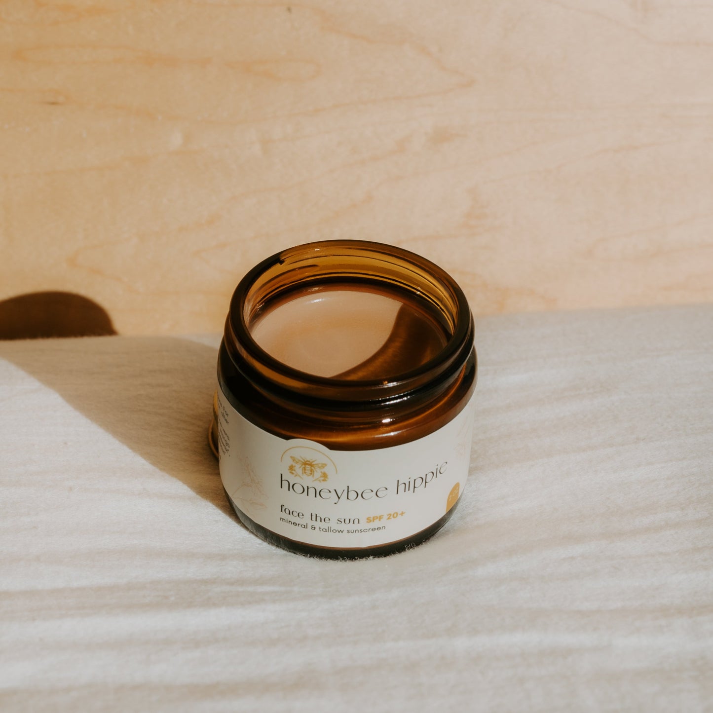 An open amber jar of Honeybee Hippie’s "face the sun - herbal tinted mineral & tallow skin protection" sits on a light surface, bathed in sunlight, showcasing natural UV defense with non-nano zinc and organic fats.