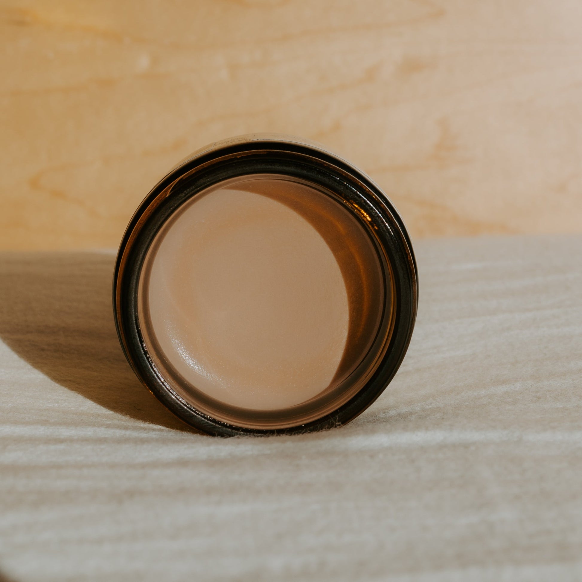 A jar of Honeybee Hippie’s face the sun—herbal tinted mineral & tallow skin protection—filled with a light brown balm, seen from above on a textured beige surface against a softly lit background.
