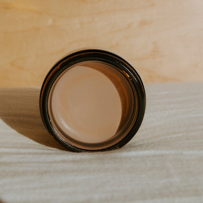 A jar of Honeybee Hippie’s face the sun—herbal tinted mineral & tallow skin protection—filled with a light brown balm, seen from above on a textured beige surface against a softly lit background.