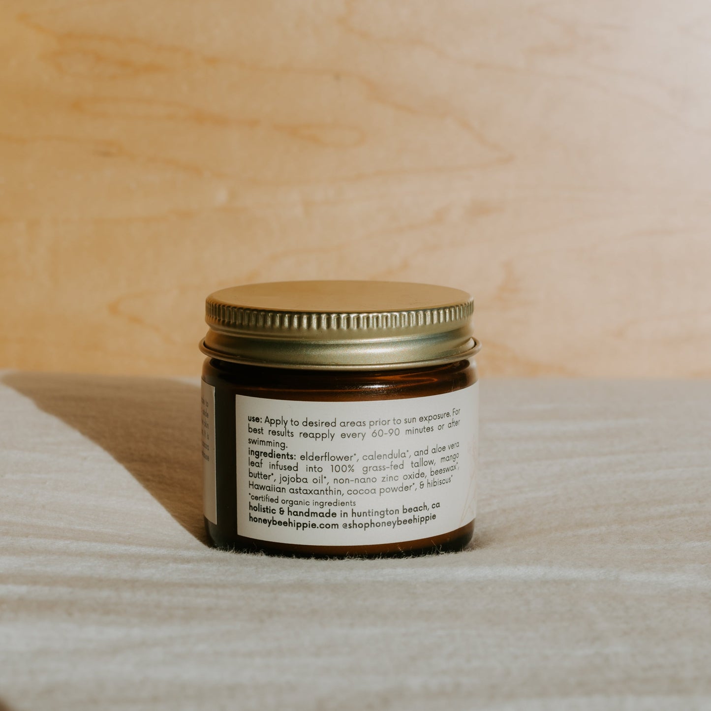 A small amber glass jar with a gold metal lid from Honeybee Hippie holds "face the sun" herbal tinted mineral & tallow skin protection, offering natural UV defense and non-comedogenic ingredients. The jar rests on a light fabric surface.