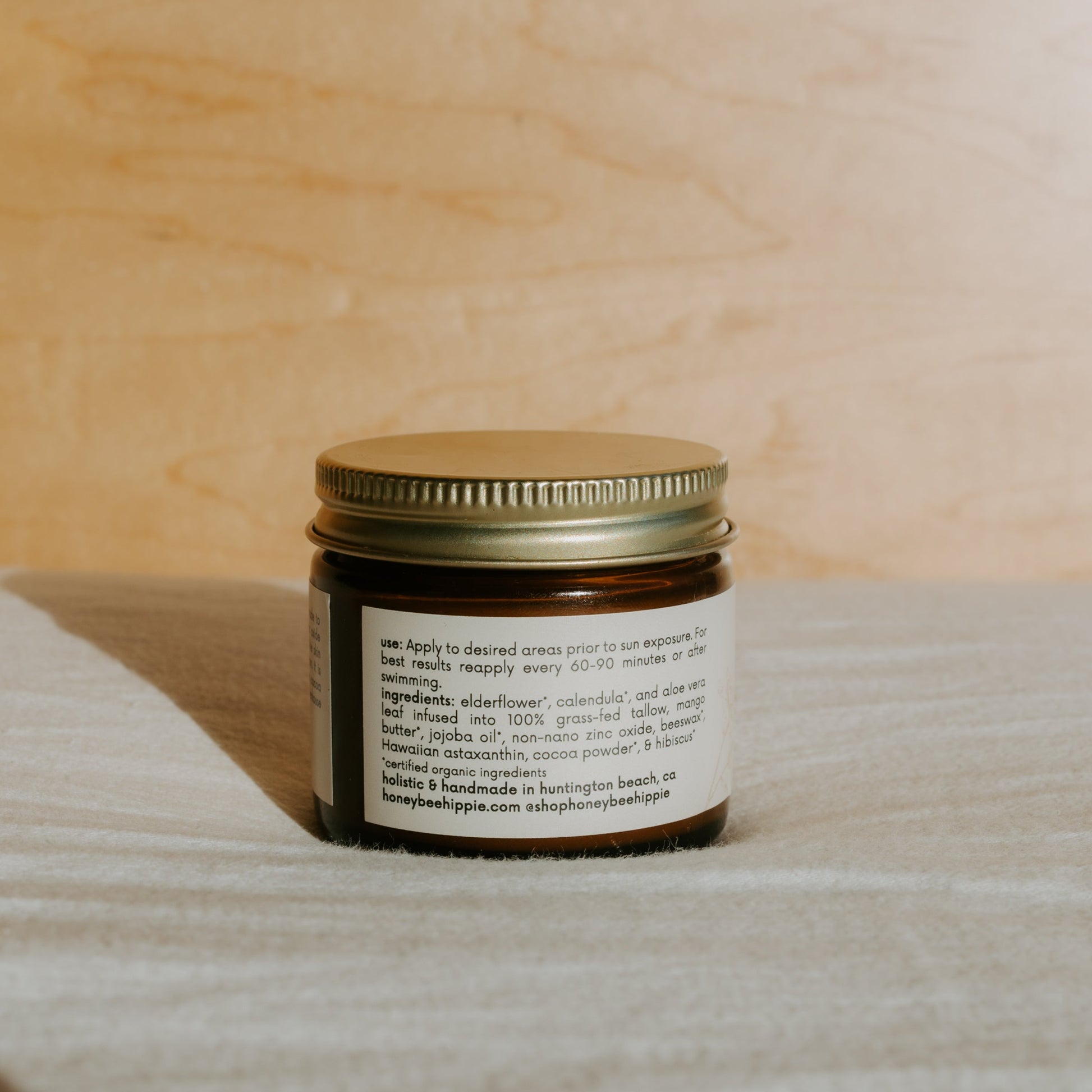 A small amber glass jar with a gold metal lid from Honeybee Hippie holds "face the sun" herbal tinted mineral & tallow skin protection, offering natural UV defense and non-comedogenic ingredients. The jar rests on a light fabric surface.
