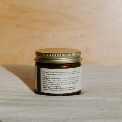 A small amber glass jar with a gold metal lid from Honeybee Hippie holds "face the sun" herbal tinted mineral & tallow skin protection, offering natural UV defense and non-comedogenic ingredients. The jar rests on a light fabric surface.