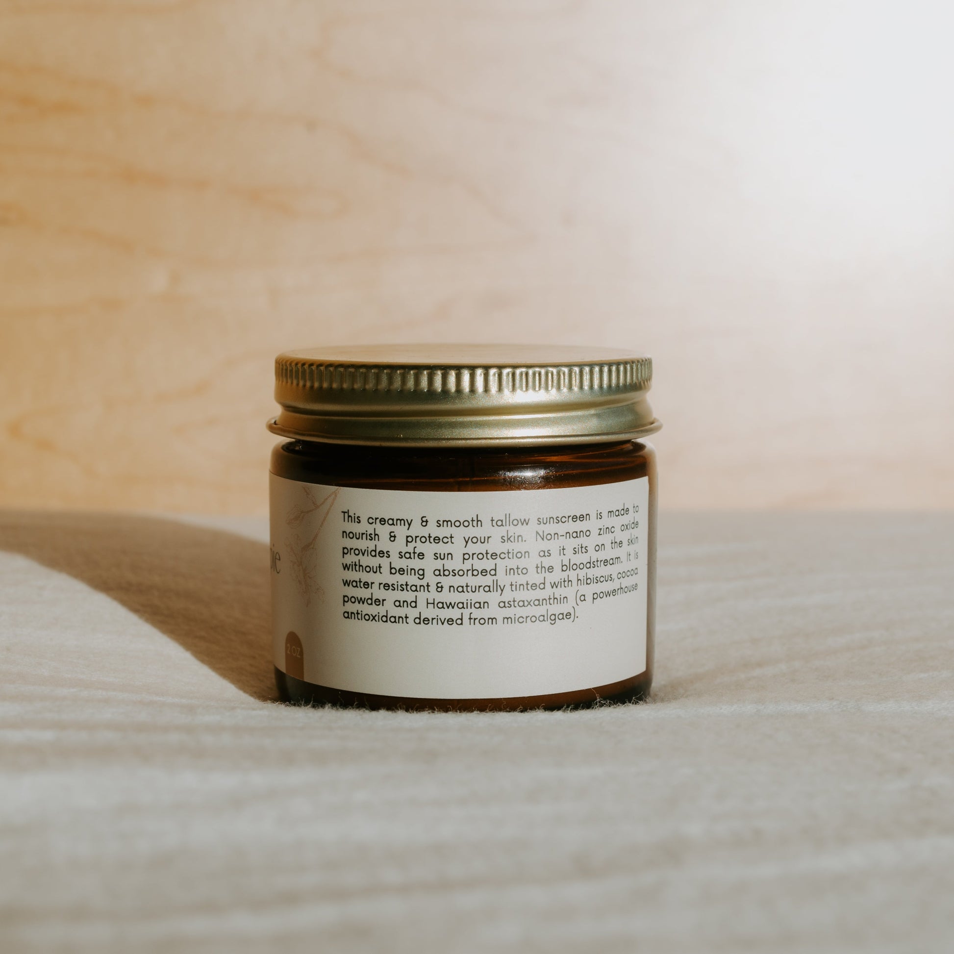 A brown glass jar with a gold lid sits on a textured surface. The label reads Honeybee Hippie face the sun—herbal tinted mineral & tallow skin protection. Sunlight casts a shadow behind the jar.