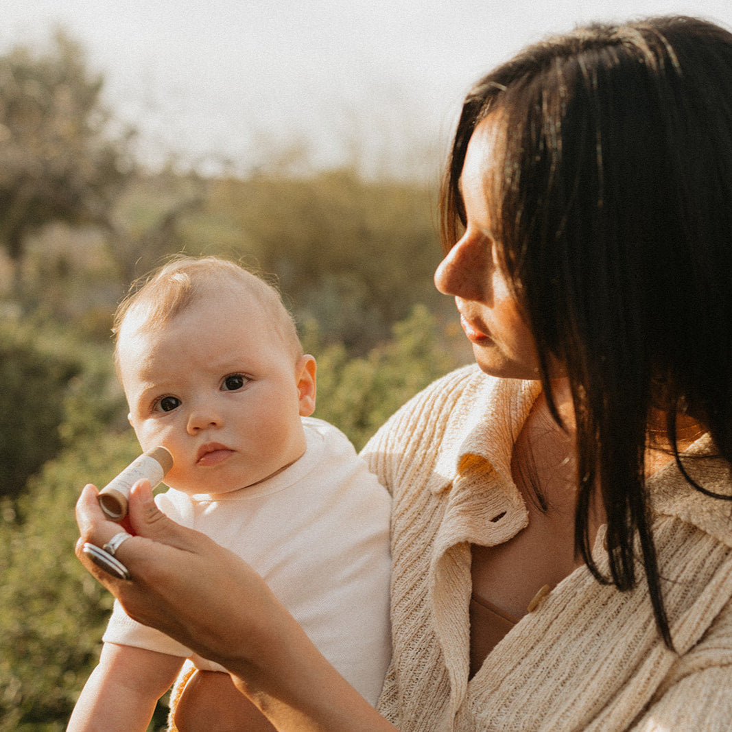 A woman gently applies Honeybee Hippie's vanilla & chamomile tallow lip butter to a baby’s cheeks in a sunlit, green outdoor setting. The baby is dressed in white, while the woman wears a light-colored sweater.