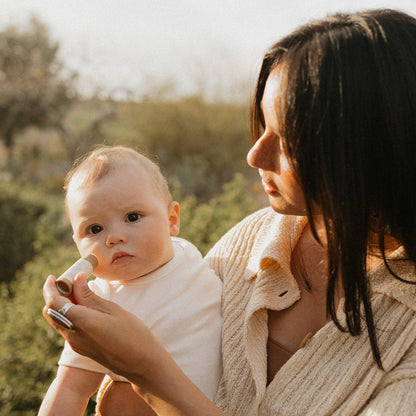 A woman gently applies Honeybee Hippie's vanilla & chamomile tallow lip butter to a baby’s cheeks in a sunlit, green outdoor setting. The baby is dressed in white, while the woman wears a light-colored sweater.