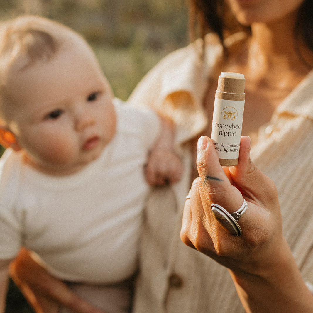 A person holds a tube of Honeybee Hippie's vanilla & chamomile tallow lip butter as a curious baby in light clothing observes. The outdoor scene is bathed in warm, natural light, creating an idyllic atmosphere.