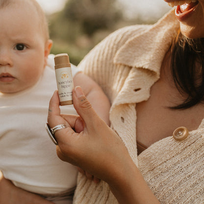 A person with a baby on their lap holds a tube labeled "vanilla & chamomile tallow lip butter" by Honeybee Hippie. Both wear light-colored clothing, set amidst soft lighting and greenery, highlighting the product.