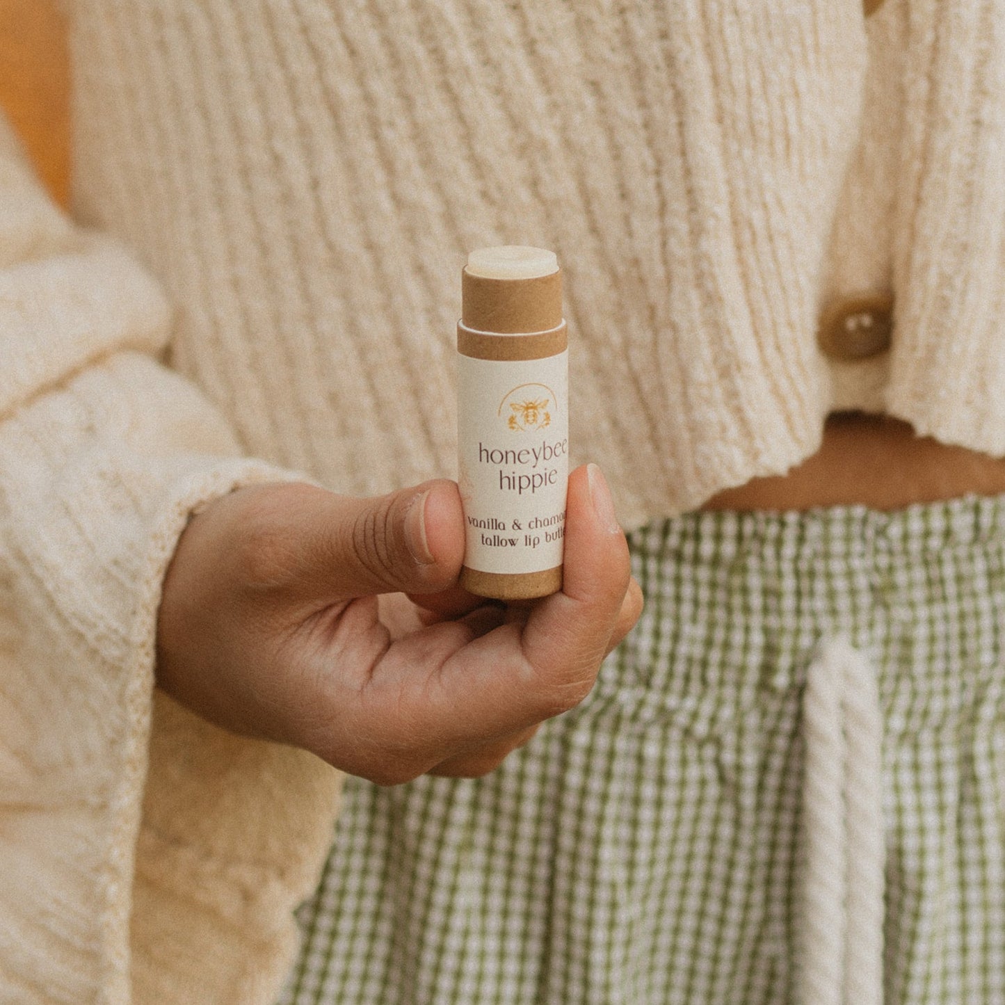 A hand holds a "vanilla & chamomile tallow lip butter" tube by Honeybee Hippie, in front of someone wearing a textured beige cardigan and green checkered pants. The softly blurred background creates a warm, cozy atmosphere.