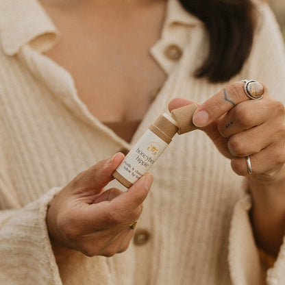 A brown-haired person with rings on their fingers, wearing a beige cardigan, holds Honeybee Hippie's vanilla & chamomile tallow lip butter in their hand. The emphasis is on them opening the tube.