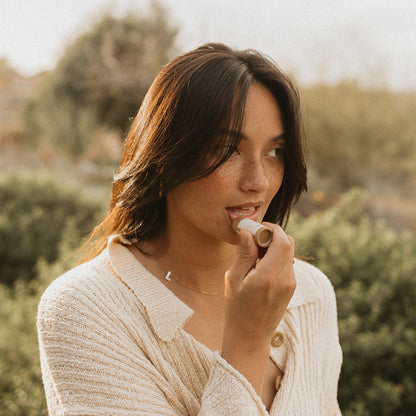 A woman with long hair and freckles applies Honeybee Hippie's vanilla & chamomile tallow lip butter outdoors, wearing a light-colored knitted sweater. The background is blurry with greenery and a hint of chamomile in the overcast sky.