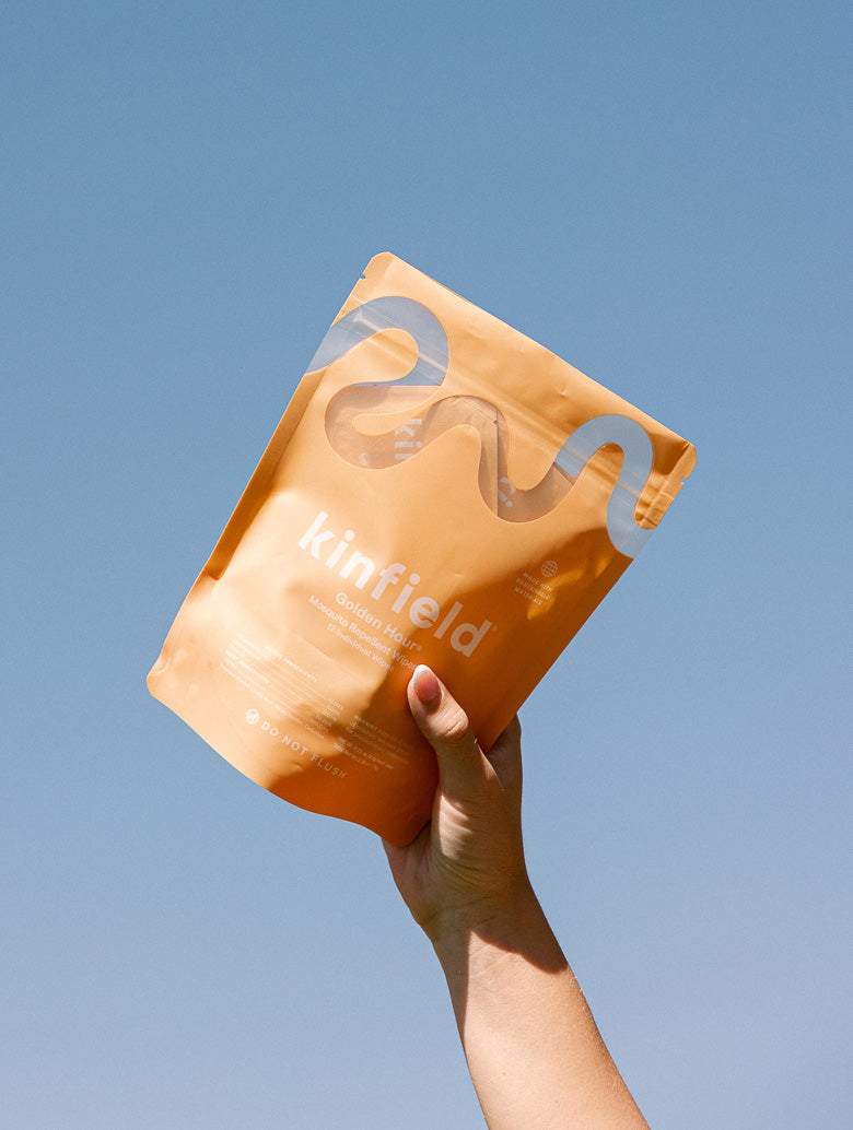 A hand holds up a bright orange, glossy pouch of Kinfield Golden Hour™ Wipes against a clear blue sky. The travel-friendly pouch features a wavy line design and minimal text.