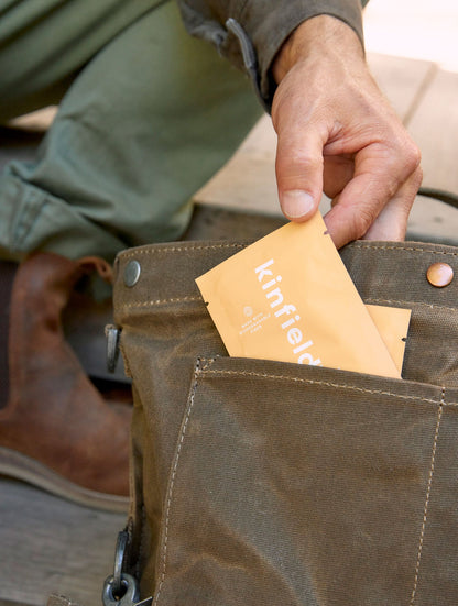 A person wearing green pants and brown boots is placing two orange packets labeled "Golden Hour™ Wipes" by Kinfield into the outer pocket of a brown canvas bag. The focus is on the person's hand and the bag containing the travel-friendly mosquito repellent wipes.