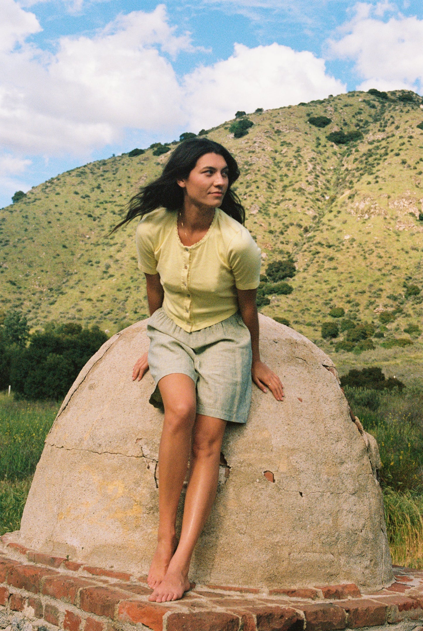A woman in a Short Sleeve Organic Cardi by LA Relaxed and a light blue skirt with lace trim sits barefoot on a large stone structure in a grassy field, gazing to her left, surrounded by green hills under a partly cloudy sky.