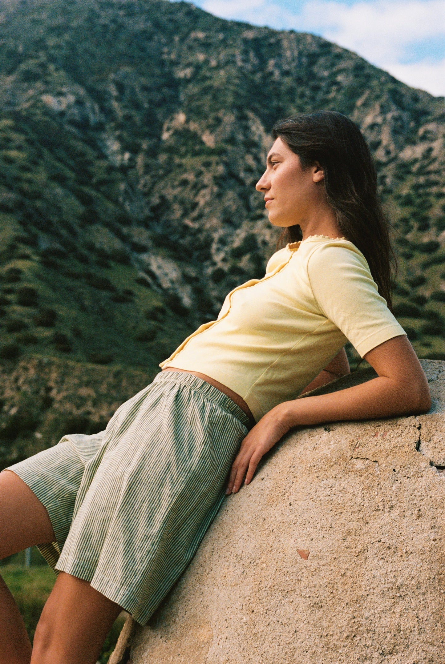 Wearing an LA Relaxed Short Sleeve Organic Cardi and striped shorts, a woman leans against a curved stone wall amidst mountainous scenery. Her long, loose hair flows as she gazes calmly into the distance, radiating serenity and reflection.