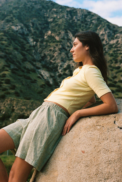 Wearing an LA Relaxed Short Sleeve Organic Cardi and striped shorts, a woman leans against a curved stone wall amidst mountainous scenery. Her long, loose hair flows as she gazes calmly into the distance, radiating serenity and reflection.