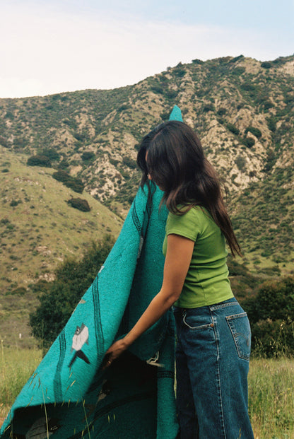 Matcha A woman stands in a grassy field by rolling hills, cradling a turquoise blanket. She wears the Bebe Organic Tee by LA Relaxed paired with a denim skirt, her dark hair cascading down. The lush hills and partly cloudy sky provide a serene backdrop to this organic cotton moment.