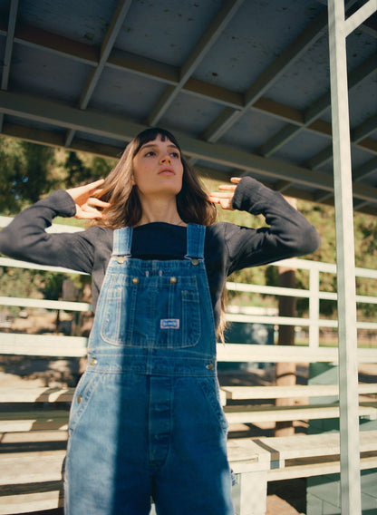 Under a metal structure on a sunny day, with trees and benches in the background, a person wearing denim overalls and an Organic Texture Long Sleeve from LA RELAXED adjusts their hair while looking upwards.