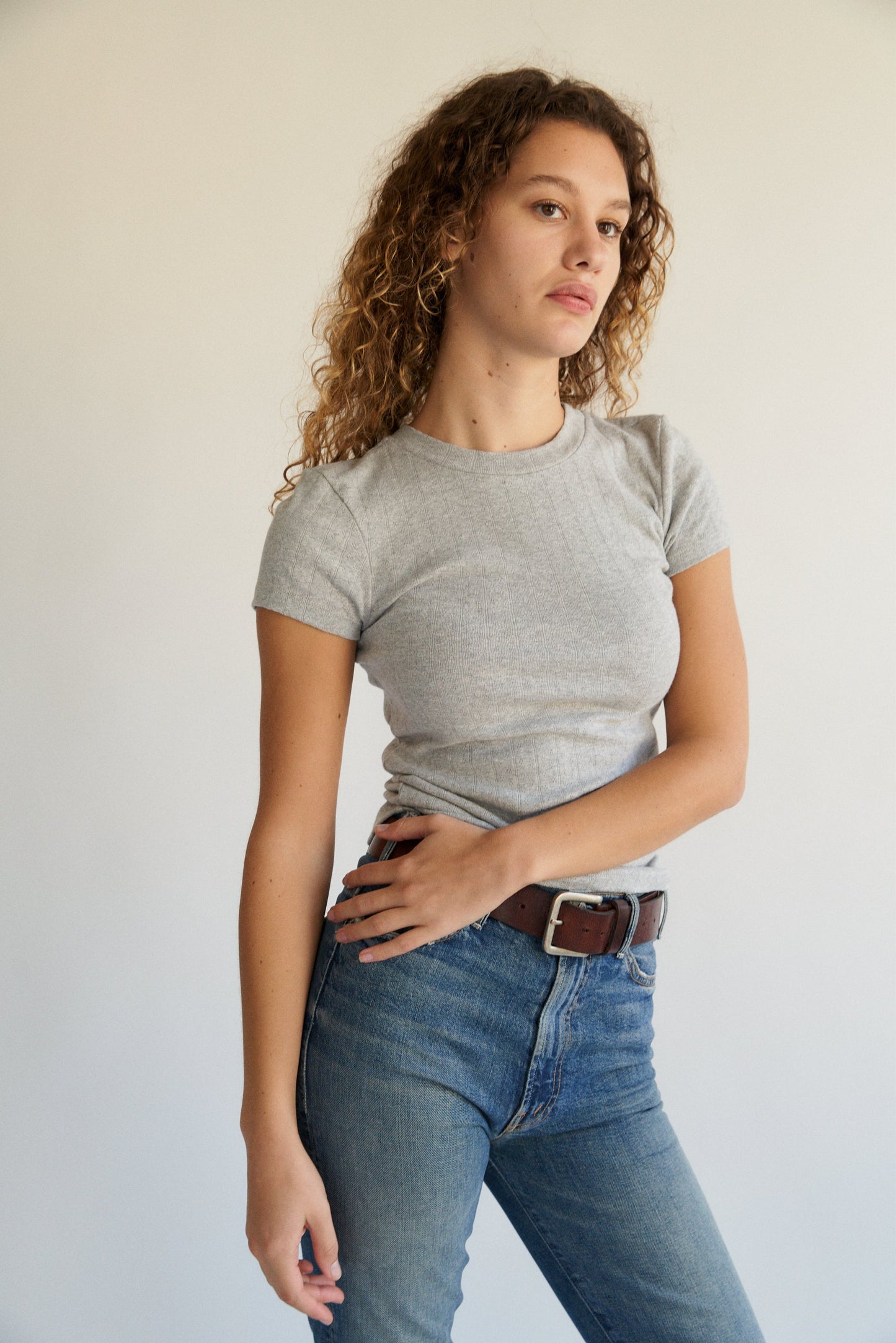 A person with curly hair is standing against a plain background, wearing an "Organic Texture Tee" by LA RELAXED in gray, paired with jeans, while looking slightly to the side in a relaxed posture.