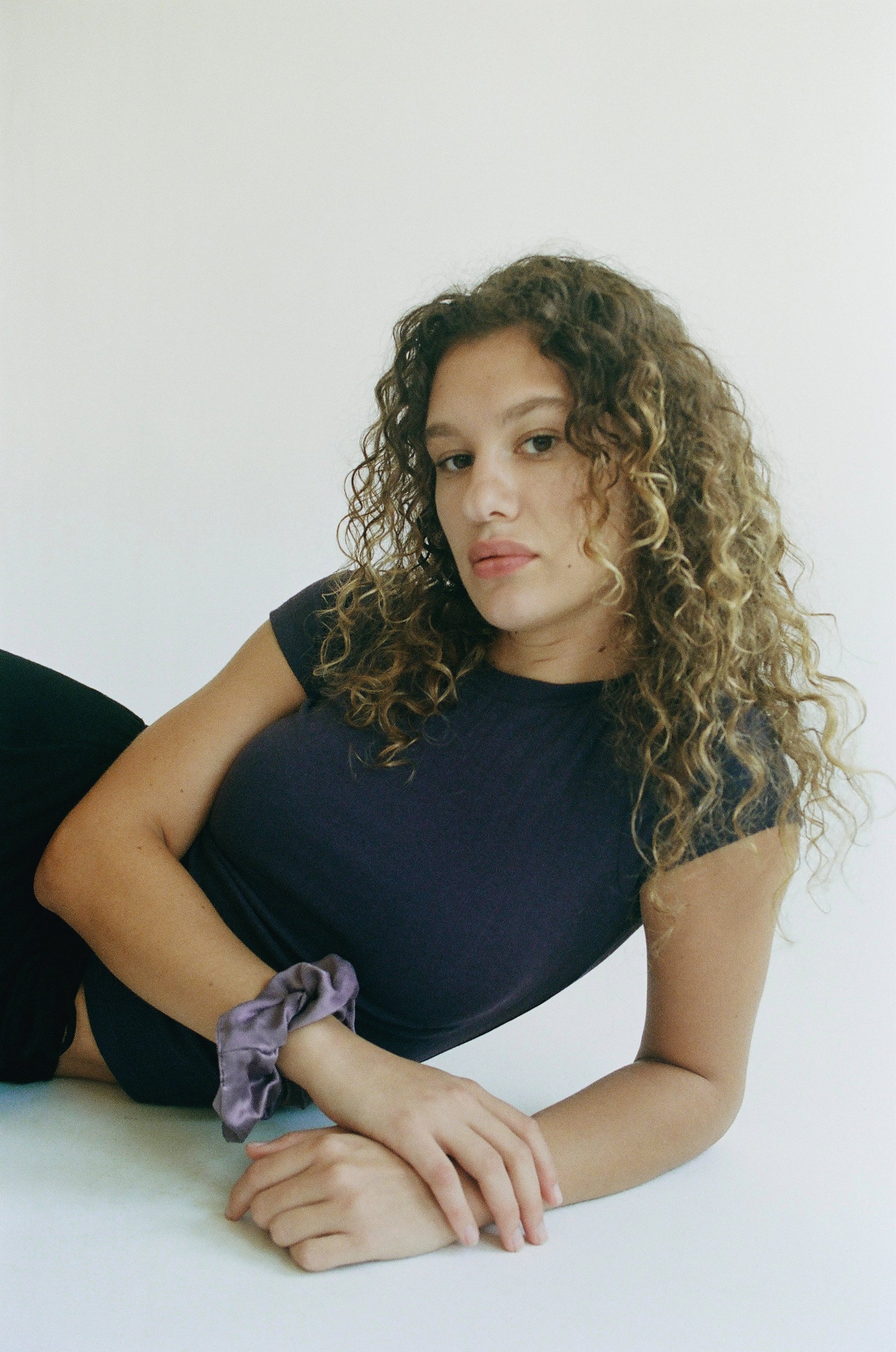 A person with curly hair lies against a white backdrop, wearing LA RELAXED's Organic Texture Tee that hints at jacquard elegance. A purple scrunchie adorns their wrist as they gaze neutrally at the camera.