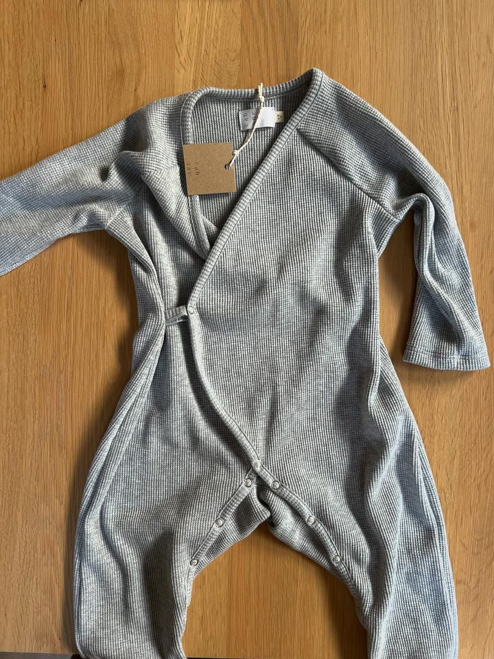 The Après Wrap Organic Waffle by cabane childrenswear is a light gray baby onesie with long sleeves and a wrap-style front, ethically handmade from organic cotton waffle fabric. It is displayed on a wooden surface with a cardboard tag at the neck.