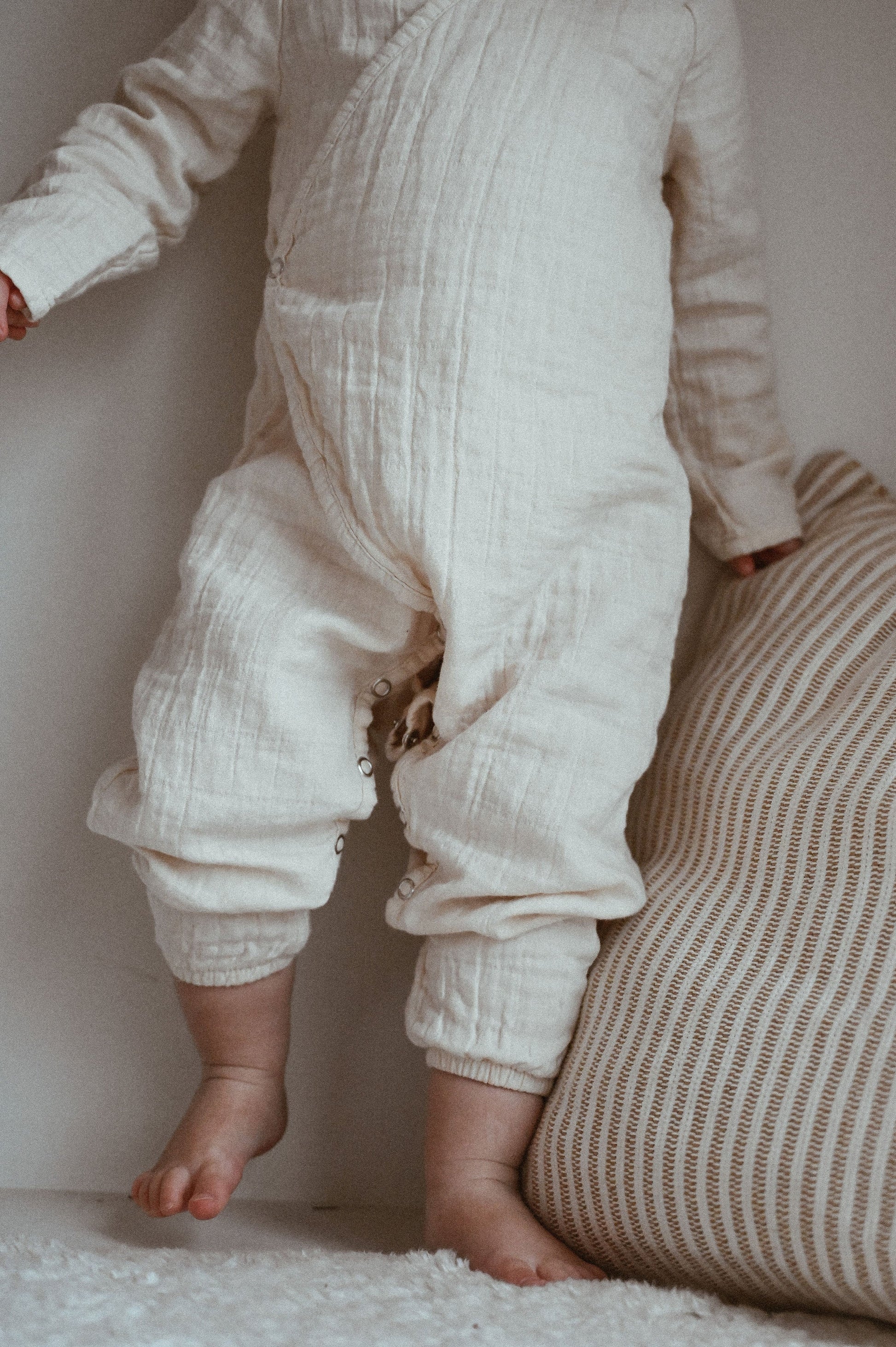 A baby wearing the cozy Après Organic Wrap Undyed from cabane childrenswear's layette collection stands on a soft surface, one hand resting on a striped cushion. Their feet are bare, and the wrap's buttons and texture are visible. 