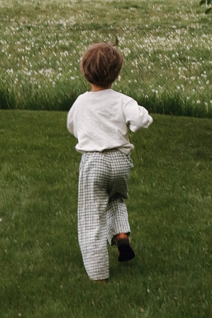 A young child with light brown hair, in a white long-sleeve shirt and cabane childrenswear PER SE PANT blue check, walks away on a green grassy lawn toward a field of white flowers.