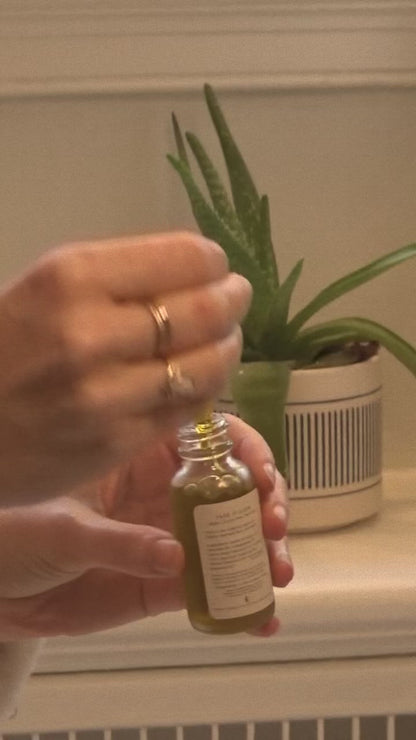 A hand holds a glass dropper above a small bottle of & Tread Well Oil Cleanser, with a potted aloe vera plant in the background on a white surface.