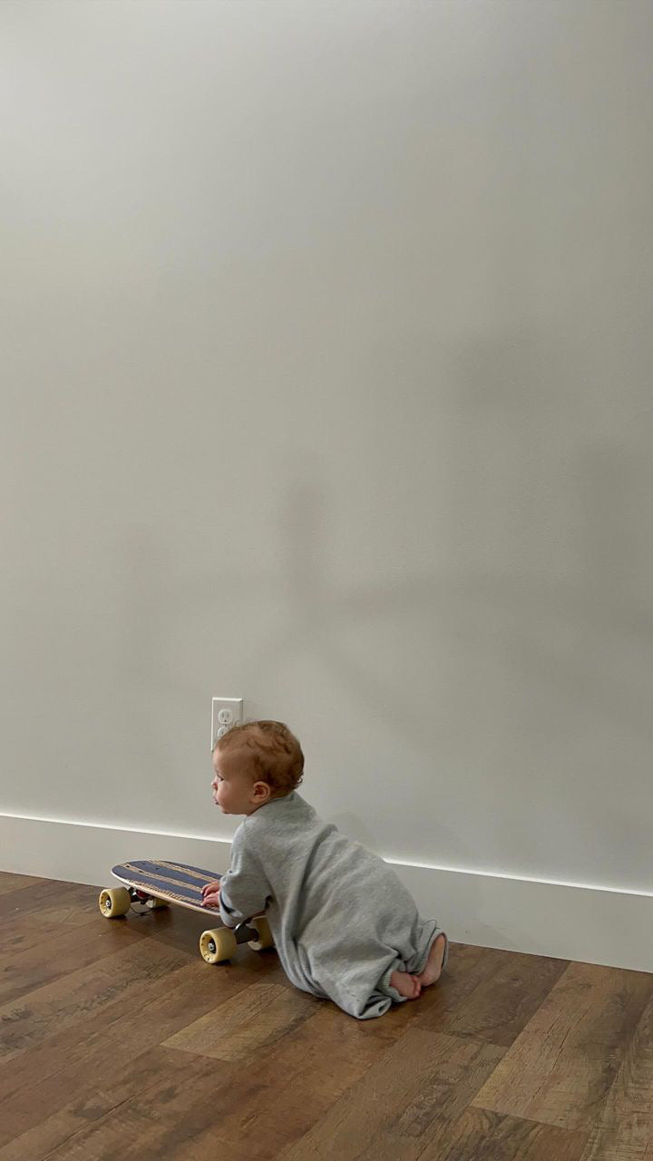 A baby in an ethically handmade gray Après Wrap Organic Waffle onesie by cabane childrenswear is kneeling on the wooden floor, clutching a small skateboard. The background is a plain light-colored wall, casting the baby's shadow.