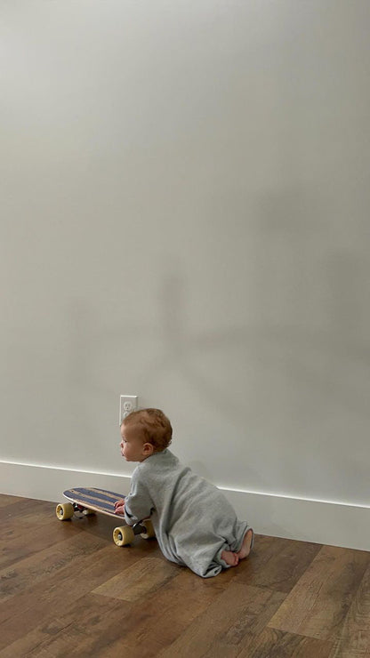 A baby in an ethically handmade gray Après Wrap Organic Waffle onesie by cabane childrenswear is kneeling on the wooden floor, clutching a small skateboard. The background is a plain light-colored wall, casting the baby's shadow.