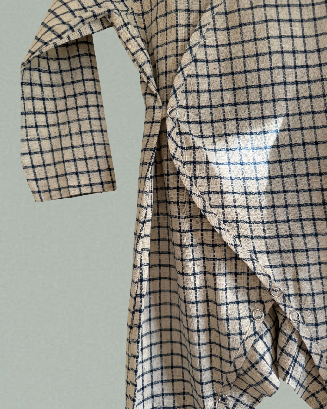 A close-up of the APRÈS WRAP blue check by cabane childrenswear, an ethically handmade baby shirt with navy blue and beige checks, side snaps, and one sleeve partially extended, shown against a plain light background.