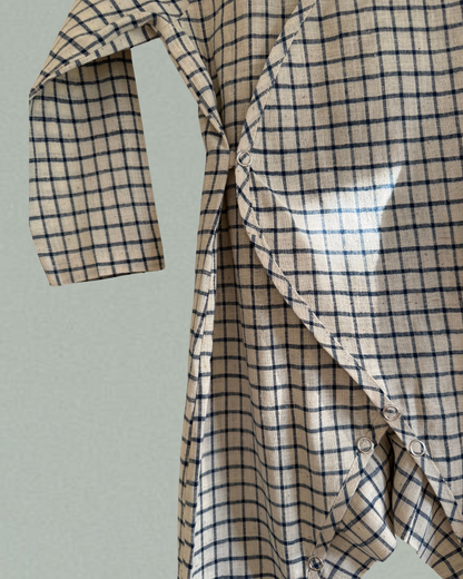 A close-up of the APRÈS WRAP blue check by cabane childrenswear, an ethically handmade baby shirt with navy blue and beige checks, side snaps, and one sleeve partially extended, shown against a plain light background.