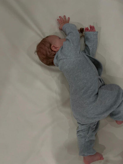 A baby in the Après Wrap Organic Waffle onesie by cabane childrenswear, made from ethically handmade organic waffle cotton, lies on a light surface sideways with curly hair, one arm reaching forward and the other stretching back.