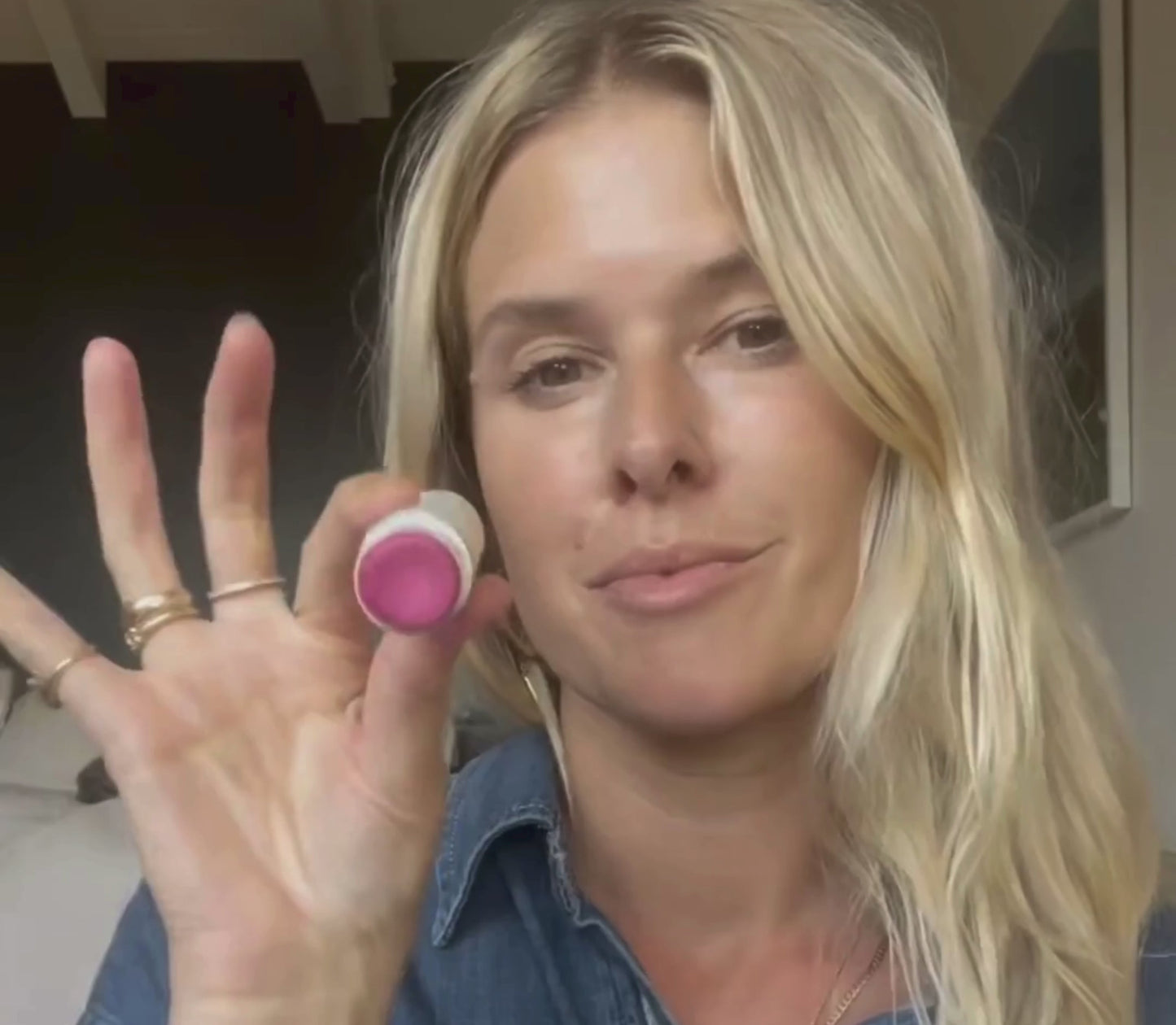 A person with long blonde hair holds a bare tint 'malibu' lip balm by Bare Essential Organics up to the camera, displaying its natural color. Dressed in a denim shirt and several rings, they stand against a blurred indoor background. The nourishing formula offers care and vibrant hues for lips and cheeks.