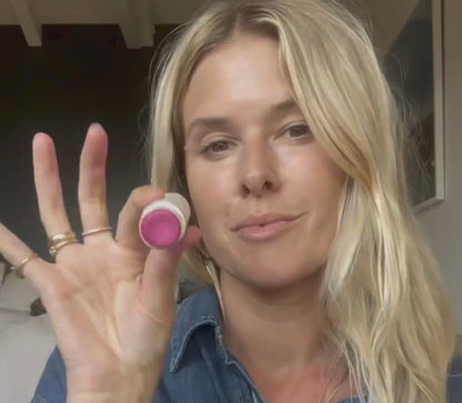 A person with long blonde hair holds a bare tint 'malibu' lip balm by Bare Essential Organics up to the camera, displaying its natural color. Dressed in a denim shirt and several rings, they stand against a blurred indoor background. The nourishing formula offers care and vibrant hues for lips and cheeks.