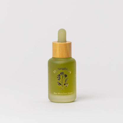 A green glass dropper bottle with a wooden cap, labeled "Blue Moon Face Serum" by My Neighbor’s Tallow and adorned with a floral motif, sits on a white background—an ideal organic serum for sensitive skin.