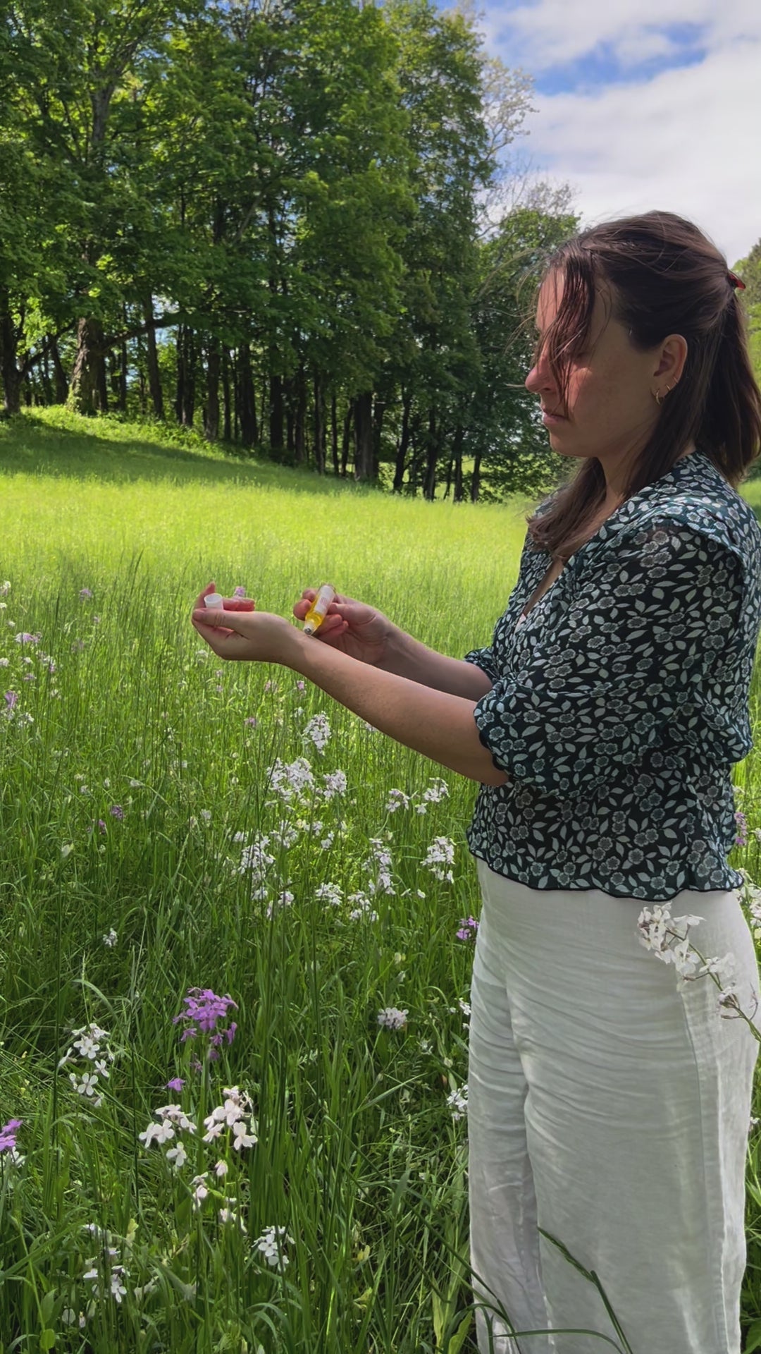 A woman in a patterned dark blouse and white pants stands in a sunlit meadow, surrounded by trees and lush grass, enjoying the gentle floral scent of & Tread Well’s Natural Fragrance – Meadow.