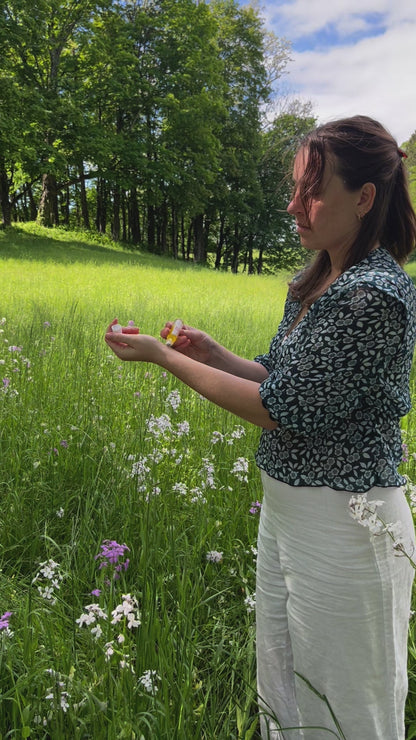 A woman in a patterned dark blouse and white pants stands in a sunlit meadow, surrounded by trees and lush grass, enjoying the gentle floral scent of & Tread Well’s Natural Fragrance – Meadow.